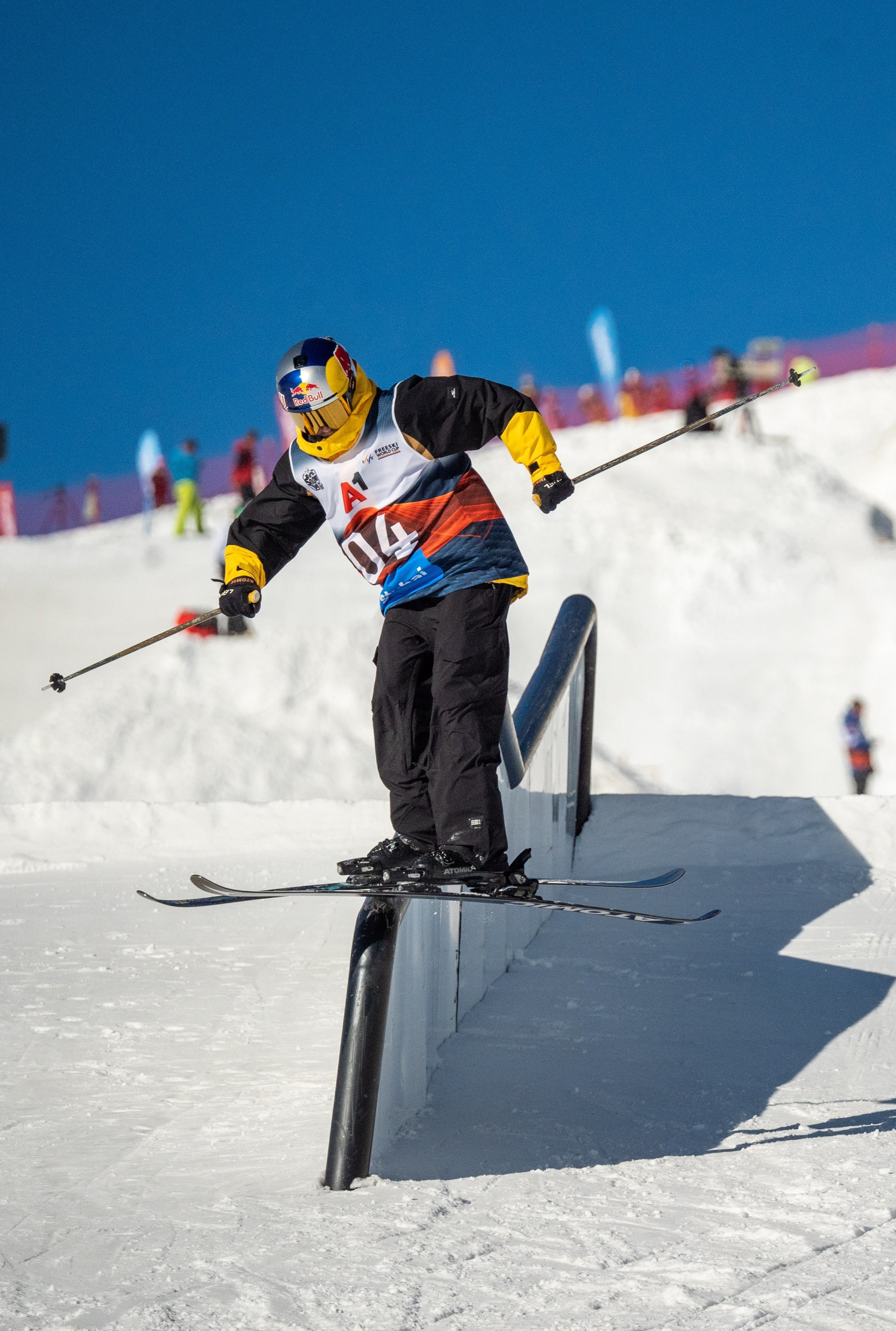 Fabian Boesch in a black, white, and yellow racing suit jumps over a metal rail on a snowy mountain under a clear blue sky.