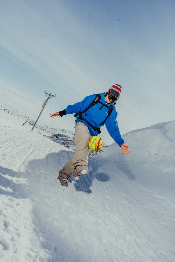 A skier in a bright blue jacket and grey pants kicking a neon yellow football mid-motion on a snowy slope, wearing a colourful striped beanie and goggles, with a wide winter landscape and pale blue sky in the background.