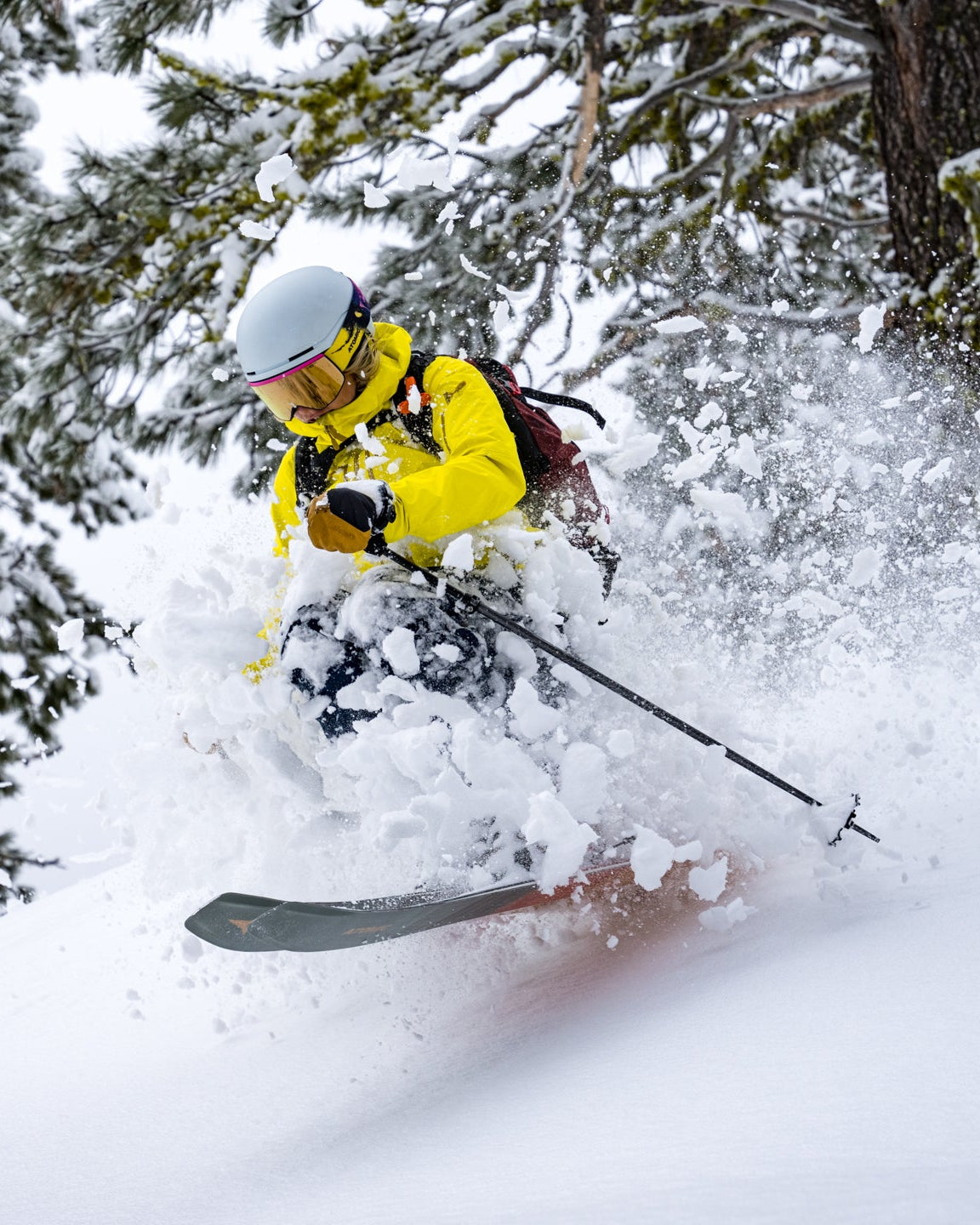 Amie Engerbretson in a bright yellow jacket, white helmet, and dark pants carves through deep powder snow, kicking up a spray of snow in a snowy forest setting.