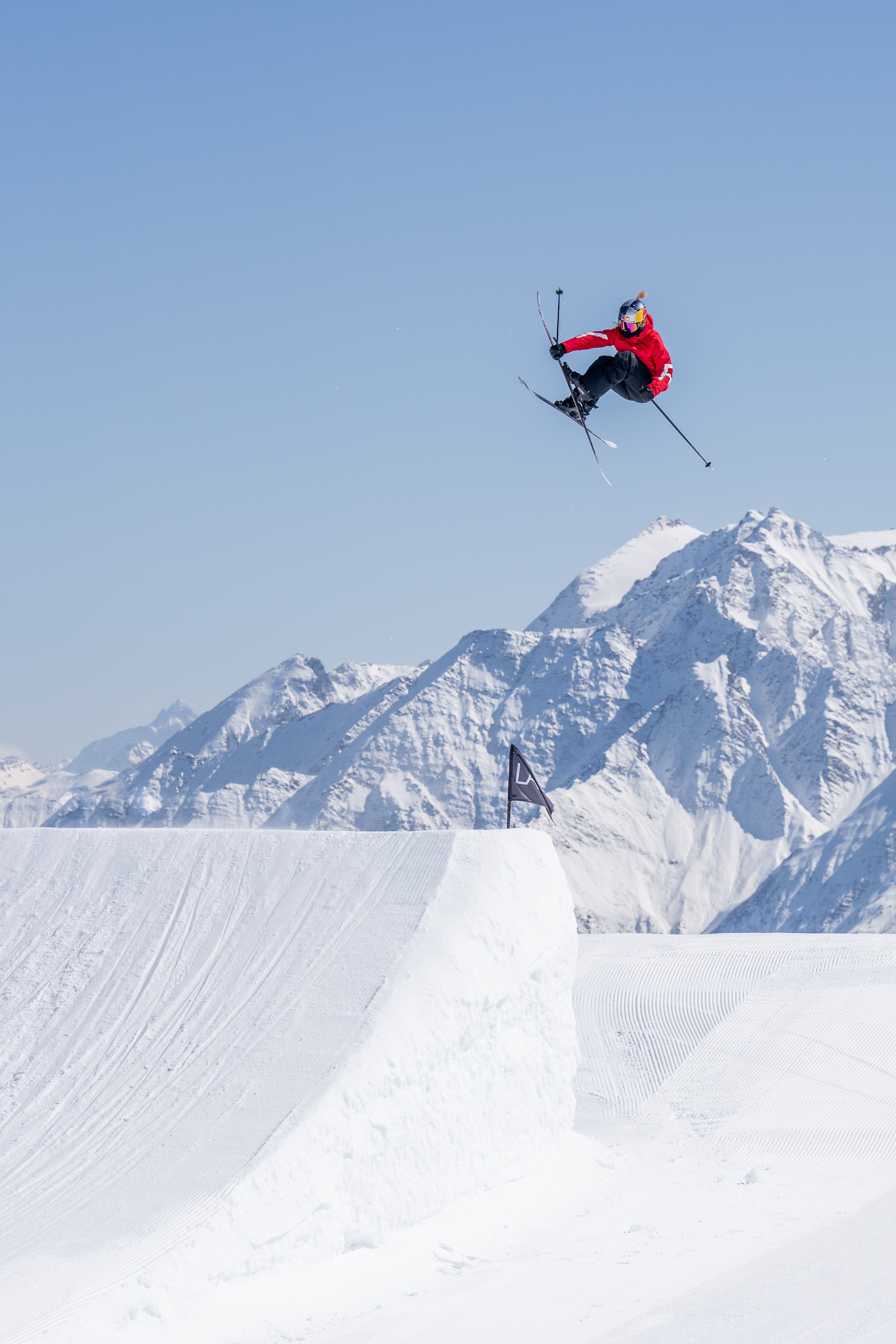 Tess Ledeux in a red jacket and black pants jumps high in the air above a snowy ramp, with snow-covered mountains and a clear blue sky in the background.