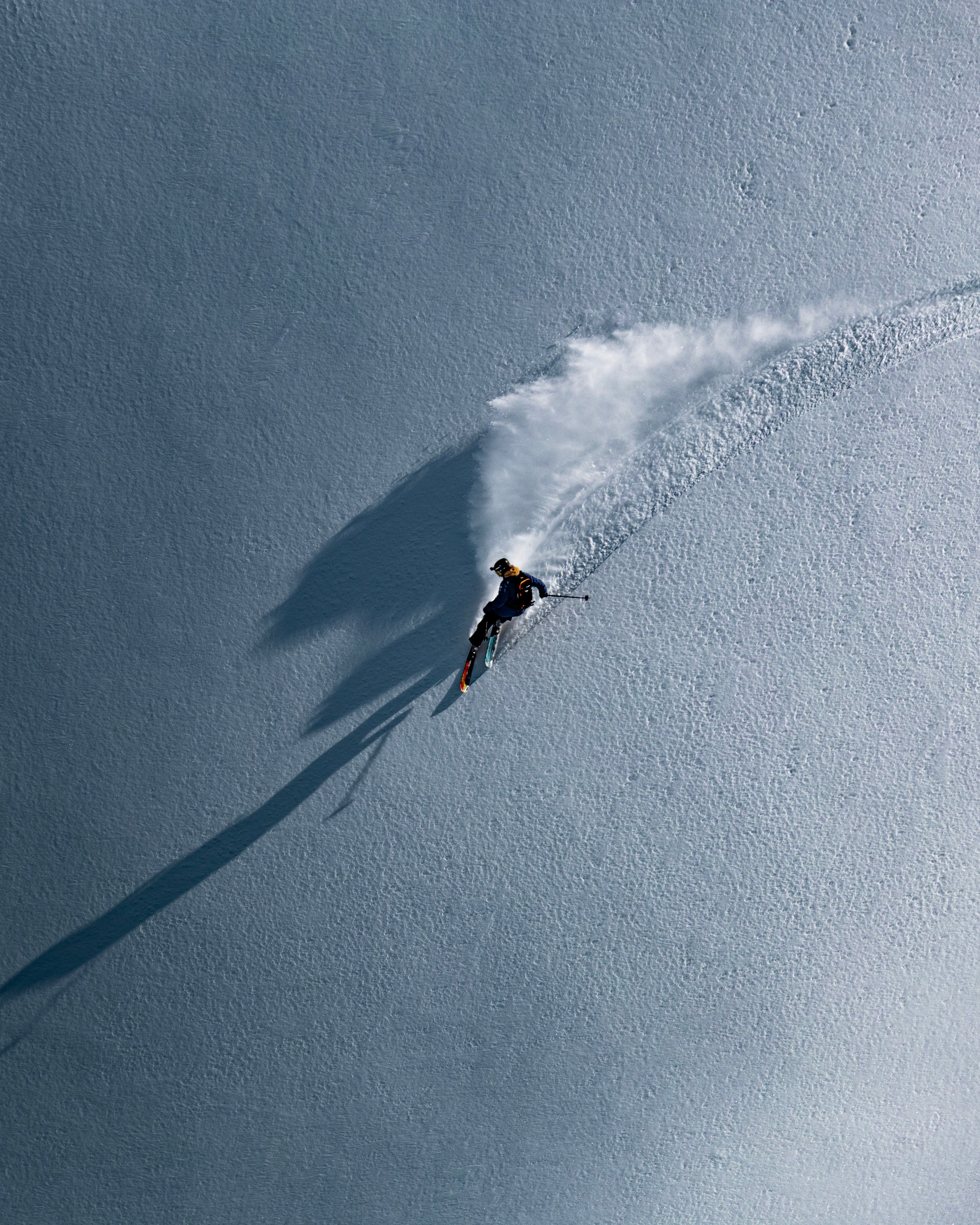 Aerial view of Nick Mcnutt carving through fresh snow, leaving a white powder trail and a long dark shadow on a bright blue-gray snowy slope.