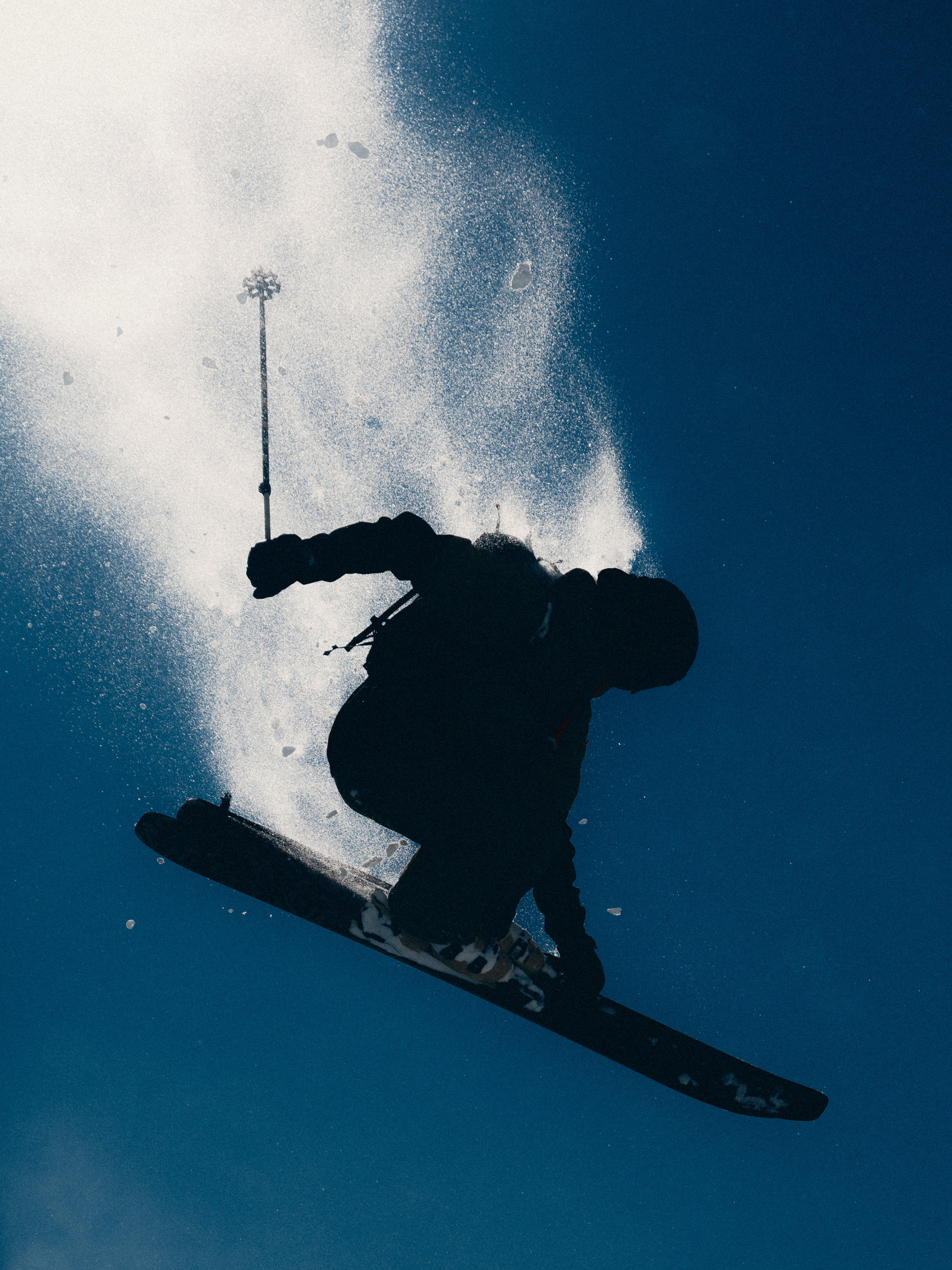 Craig Murray in dark winter gear mid-jump against a deep blue sky, with a dramatic spray of snow behind.