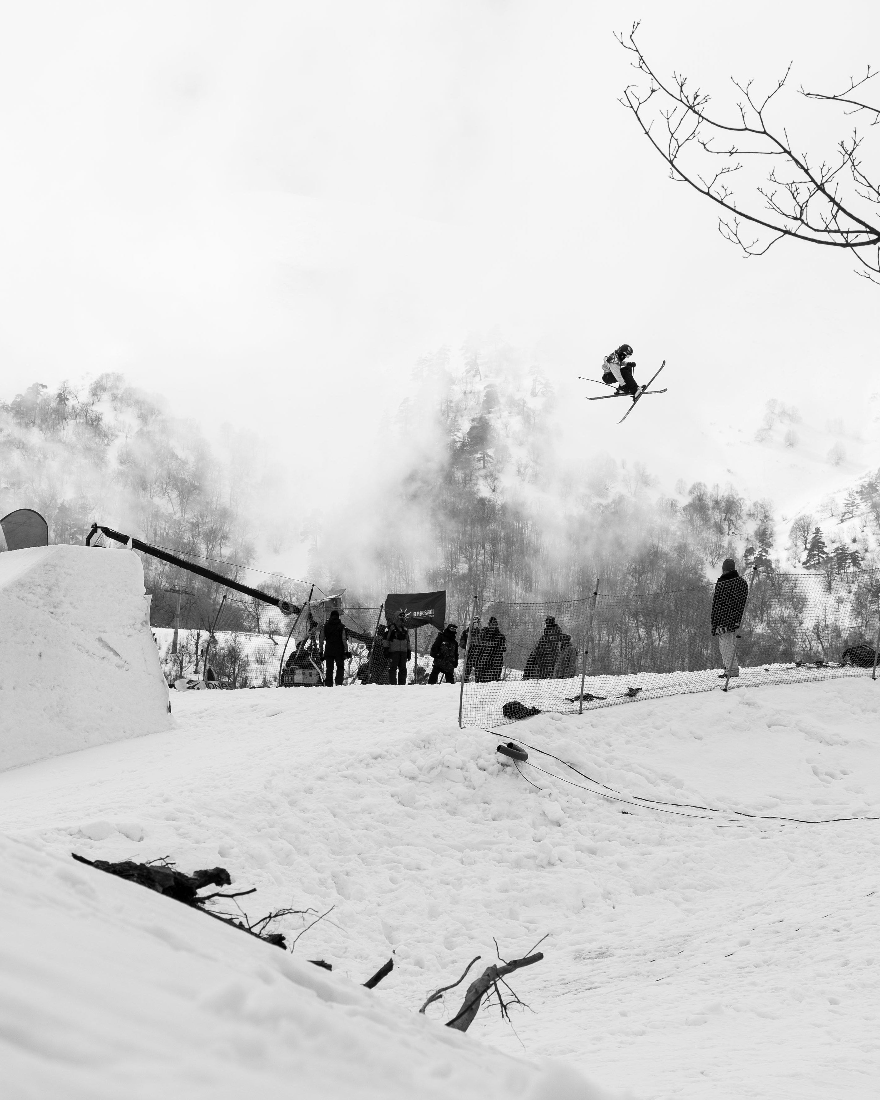 Black and white action photograph of Megan Oldham in mid-air performing a jump at a snowy mountain resort, with other skiers and snow features in the background.