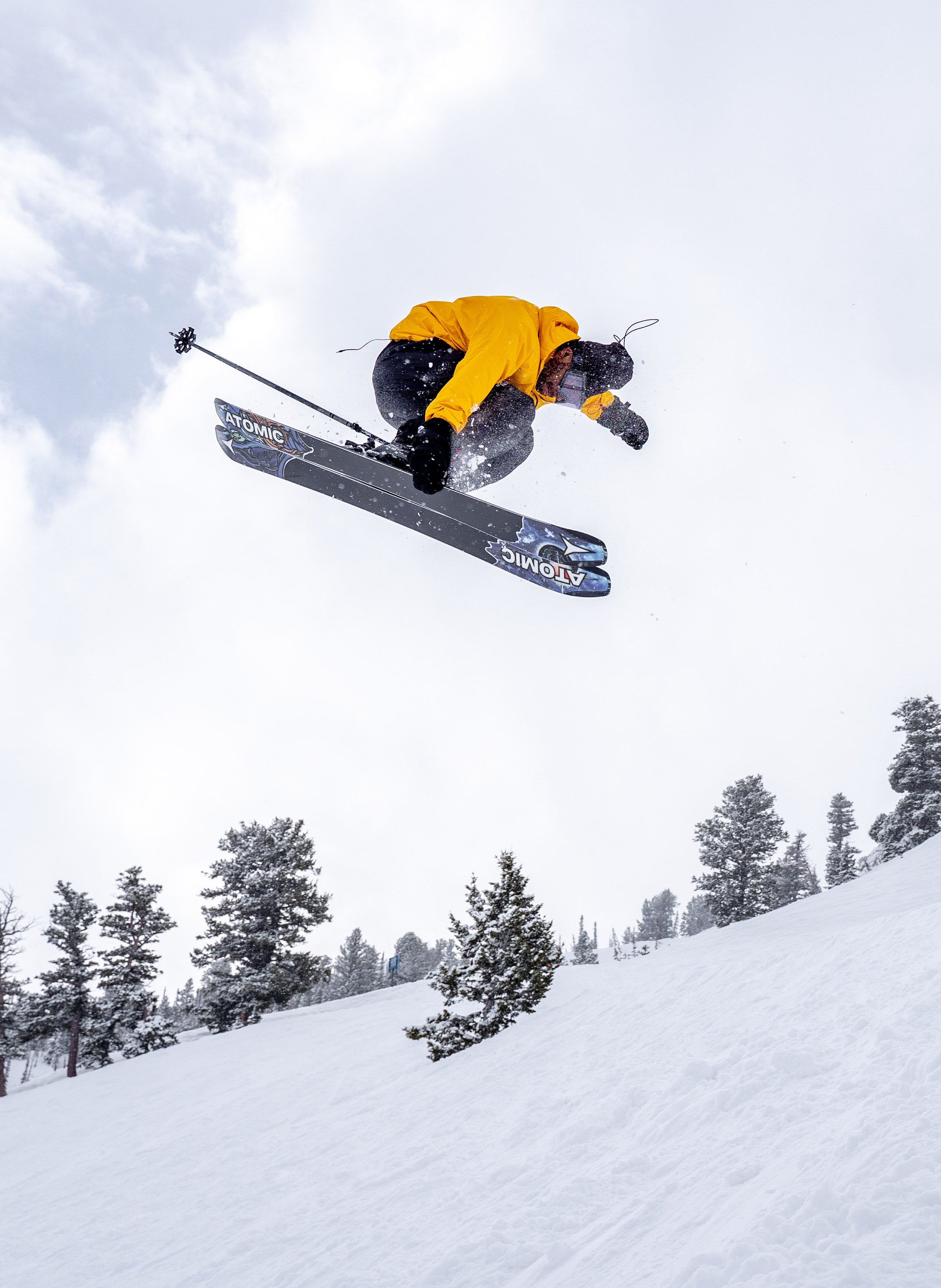 Dennis Ranalter in a bright yellow jacket and black pants performs a mid-air jump on snowboard with ski poles, set against a snowy mountain slope and pine trees under a cloudy sky.