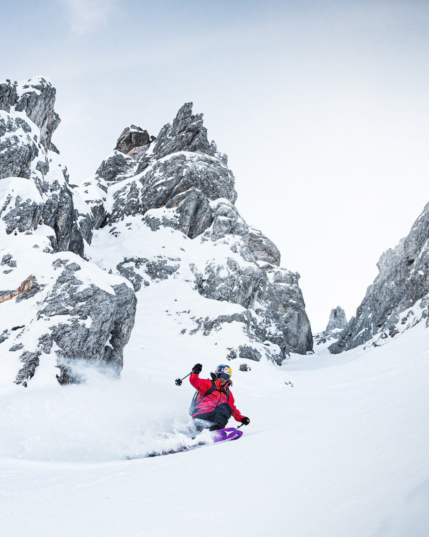 Arianna Tricomi in a bright red jacket and purple boots carving through deep powder snow between dramatic rocky mountain peaks under a pale sky.