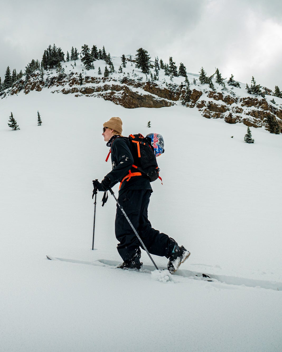 A person skiing through deep snow on a mountain slope under an overcast sky, wearing a black ski outfit, tan beanie, and a bright orange and blue backpack.