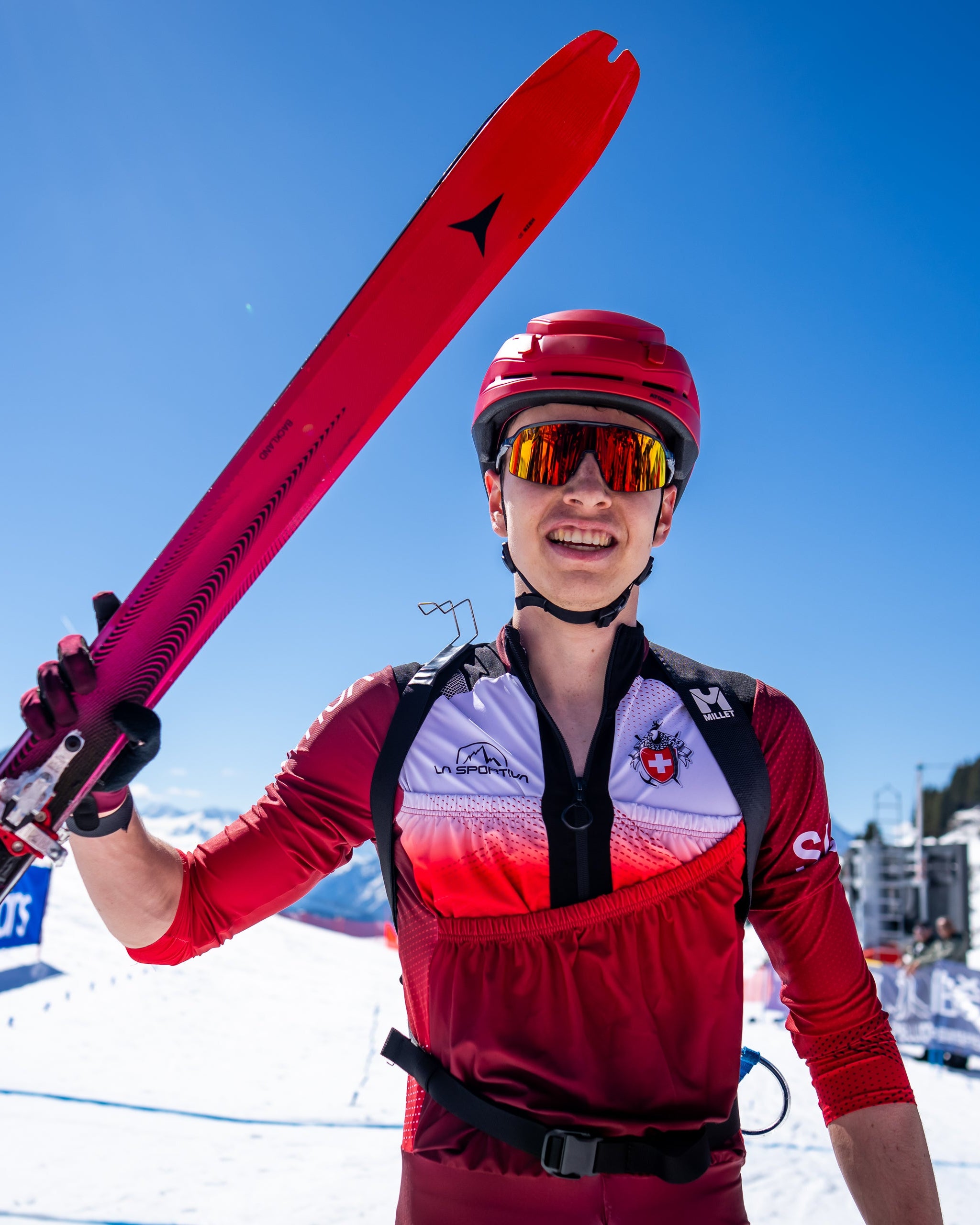 Jon Kistler wearing a red Backland UL helmet, ski goggles, and a red and white athletic jersey holds a pair of Backland 65 UL skis up against a bright blue sky at a snowy mountain event.