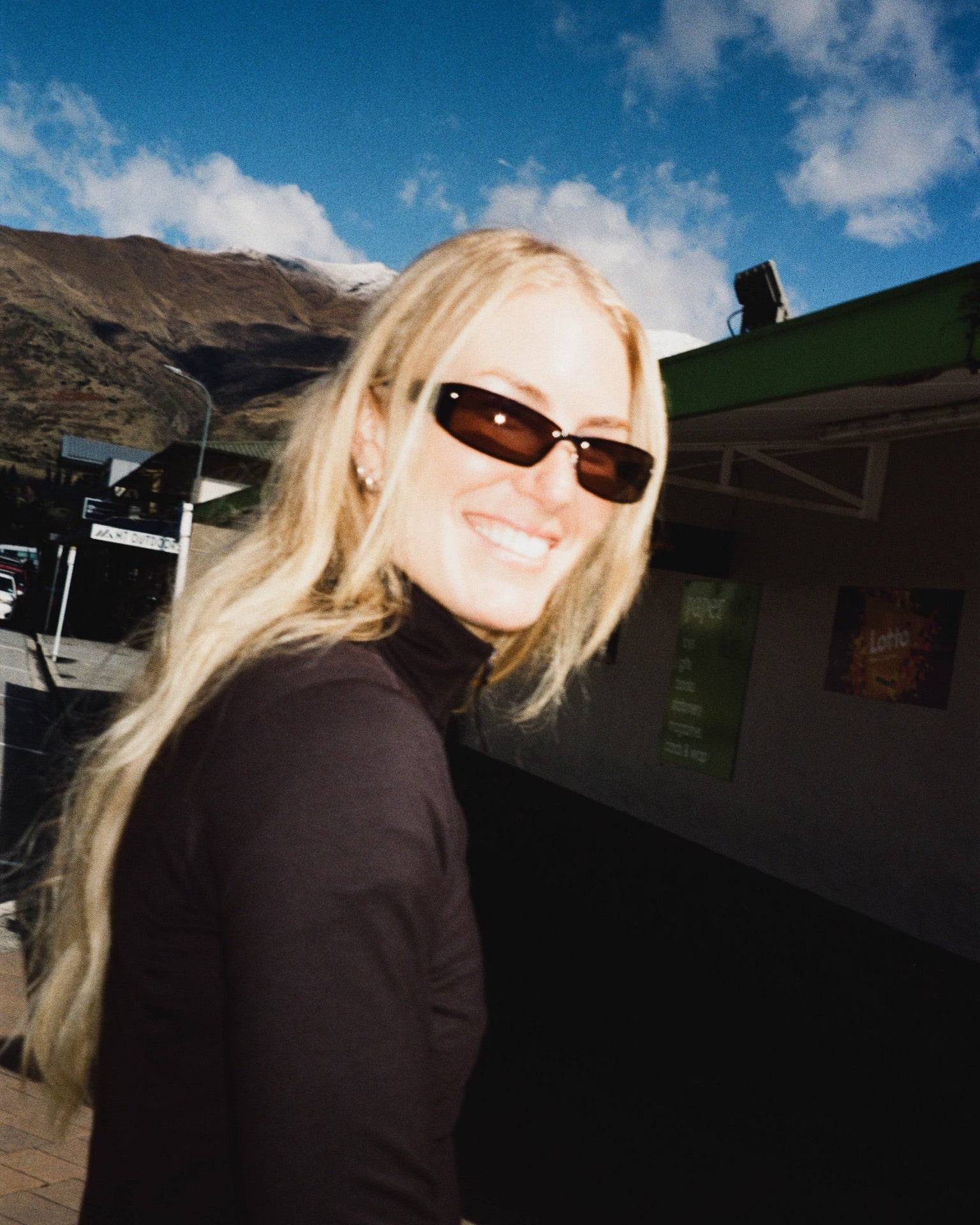 Tonje Kvikik swearing dark sunglasses and a black turtleneck top, photographed outdoors against a blue sky, mountains, and a building.