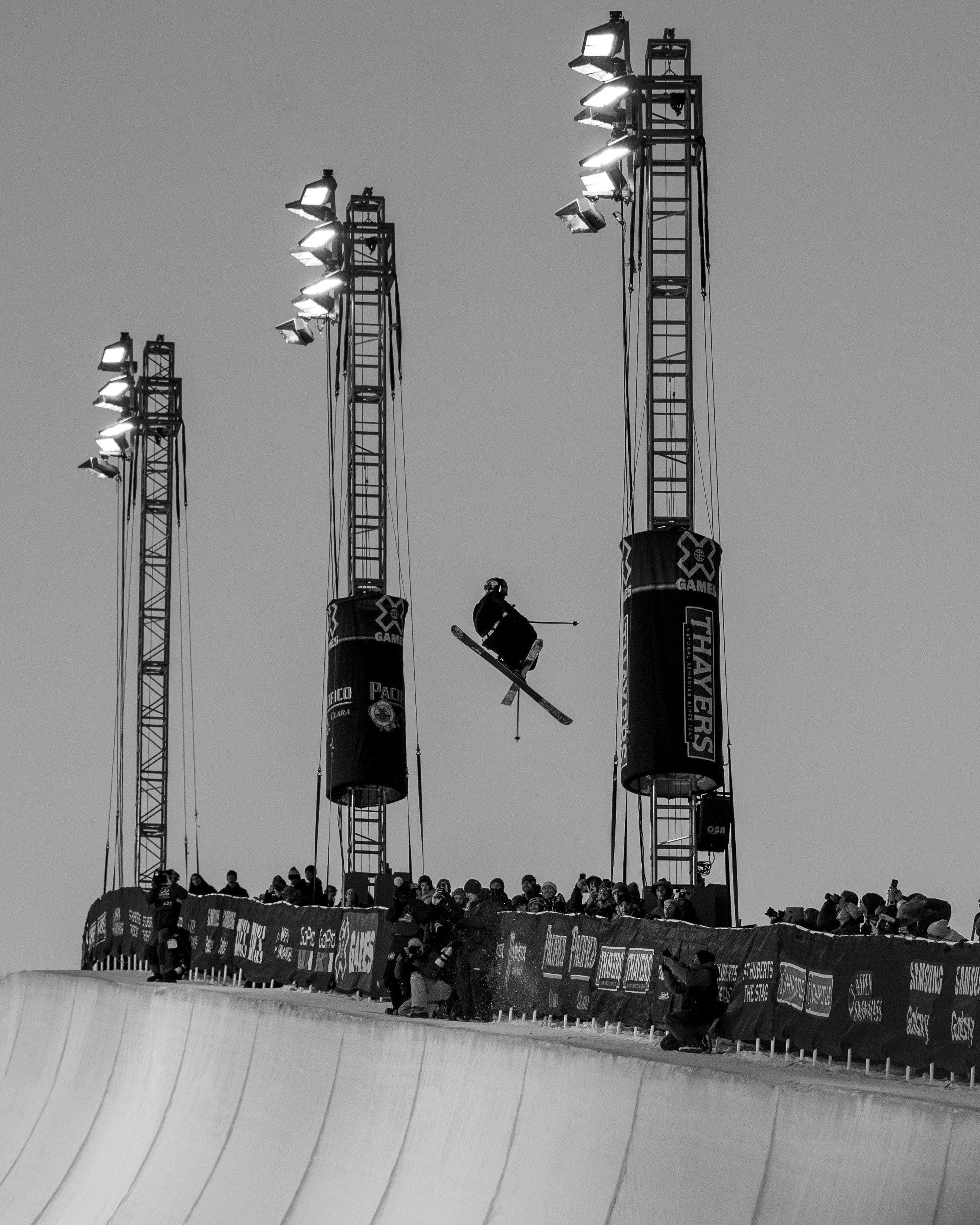 Black and white action photograph of Nico Porteous mid-air performing a trick on a large halfpipe, with tall stadium lights and a crowd of spectators in the background.