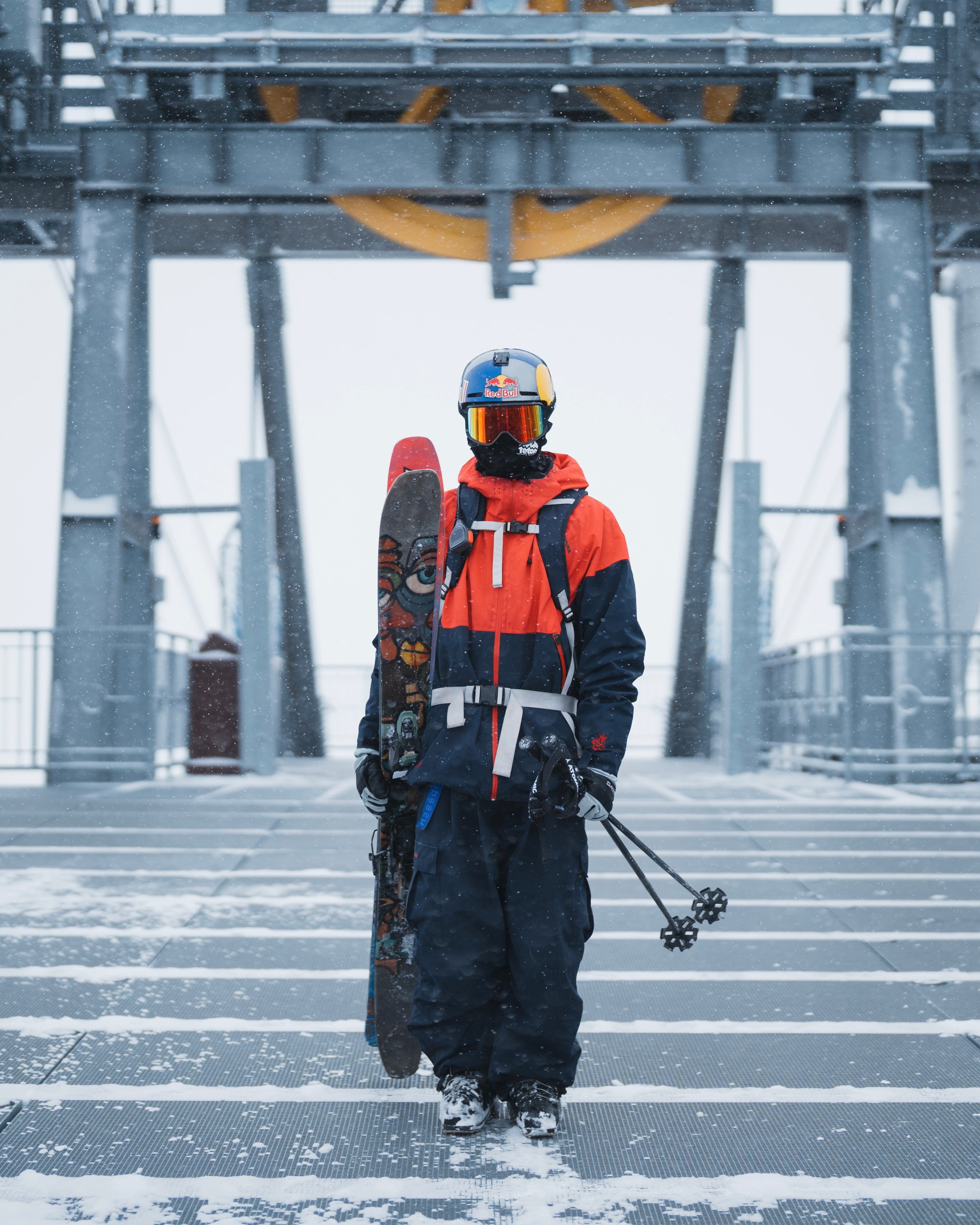 A snowboarder stands on a snowy bridge in the snow, wearing a bright orange and navy ski jacket, dark pants, a blue helmet with orange goggles, and carrying a snowboard and ski poles.