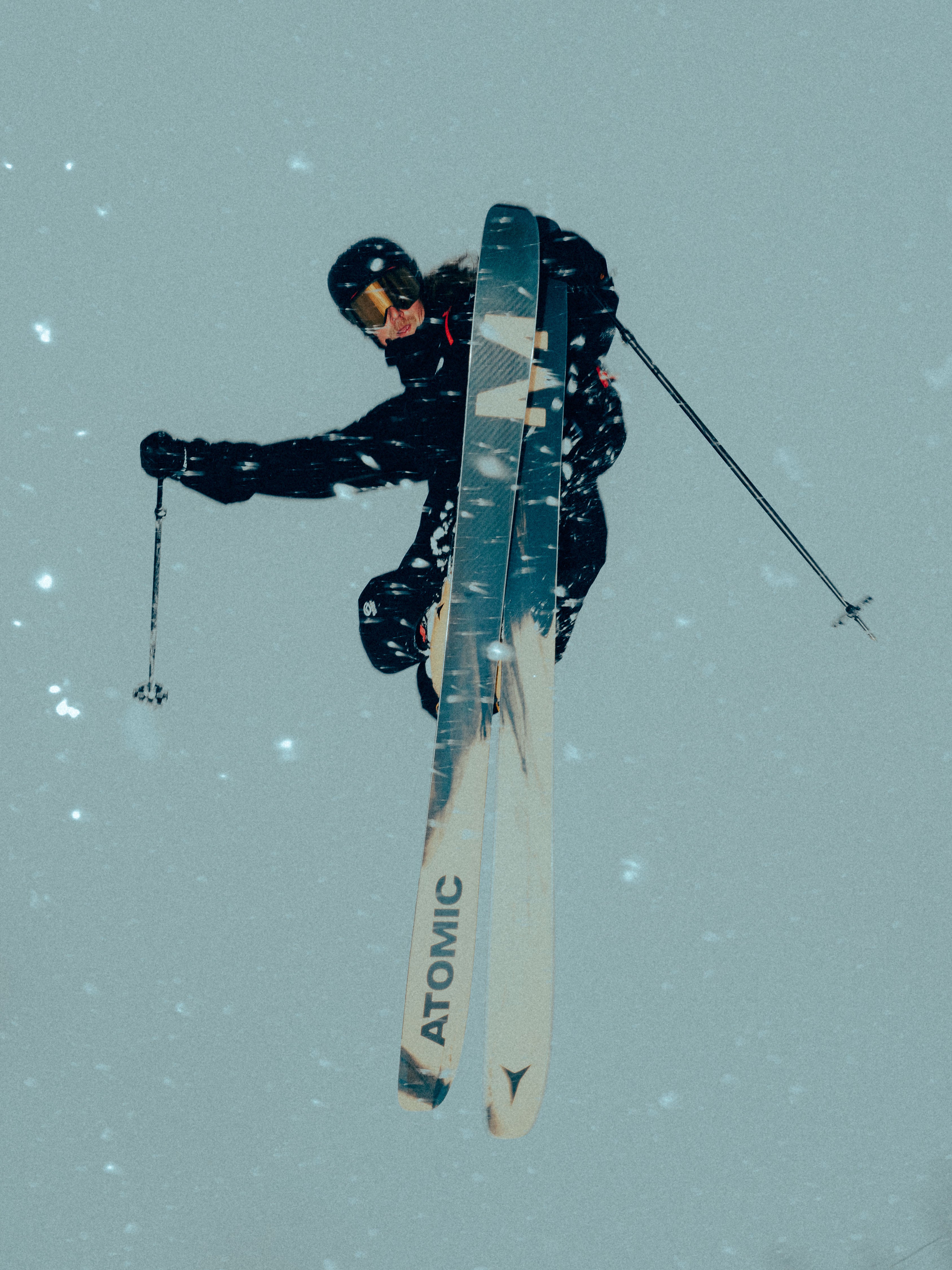 Nick Mcnutt mid-jumping in the snow, wearing a black ski suit and helmet, with Atomic skis and ski poles visible against a snowy gray-blue background.