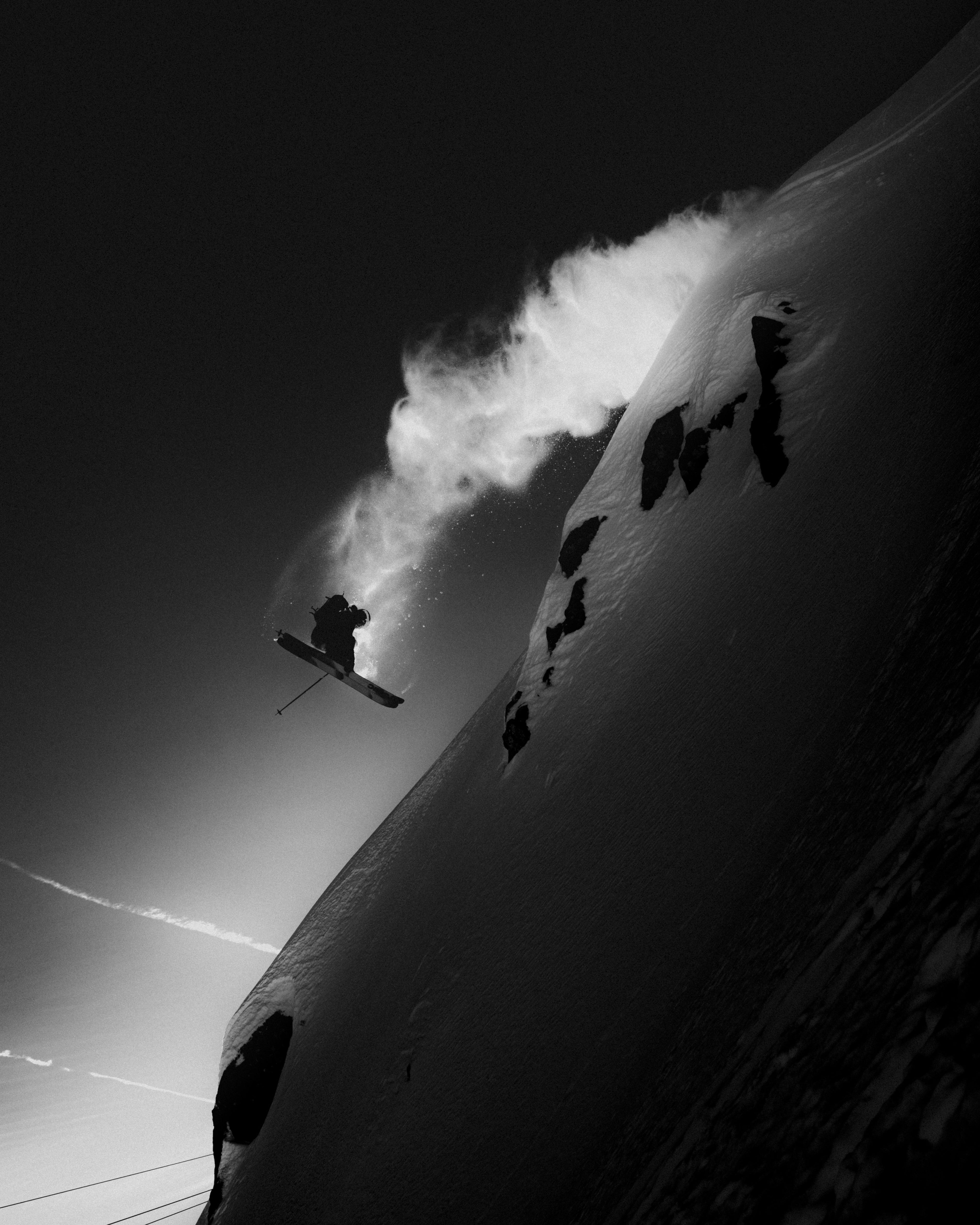 Black and white action photograph of Craig Murray mid-jumping against a snowy mountain slope, with a dramatic cloud of snow in the background.