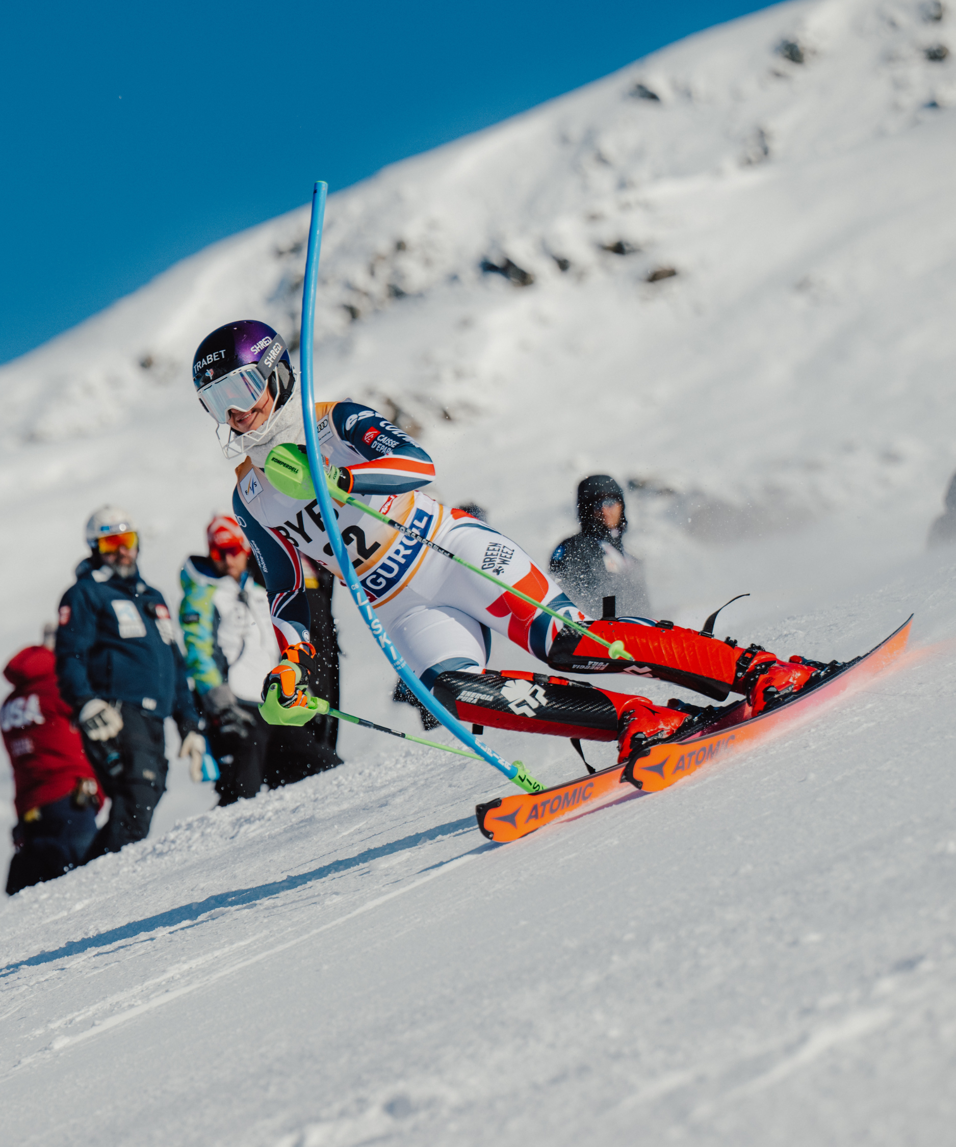 Marion Chevrier in a white, orange, and blue racing suit leans sharply into a turn on a snowy mountain slope under a clear blue sky, with spectators visible in the background.