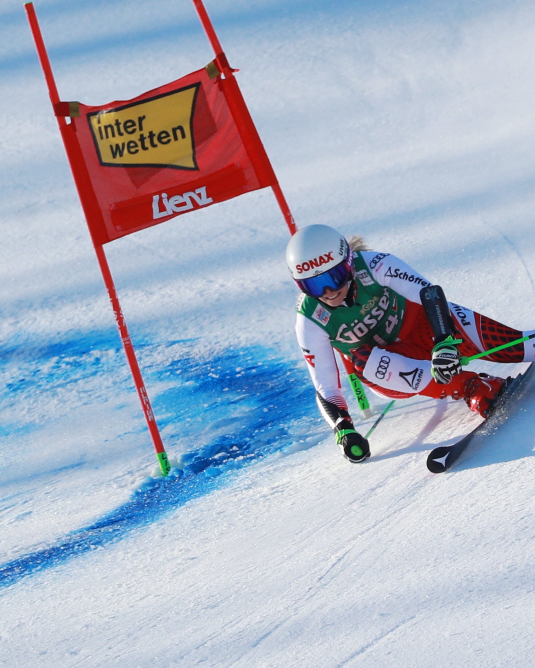 Eva Maria Braem in a red, white, and green racing suit navigates a snowy slope beside a red Interwetten-Liebhahn flag during a downhill race.