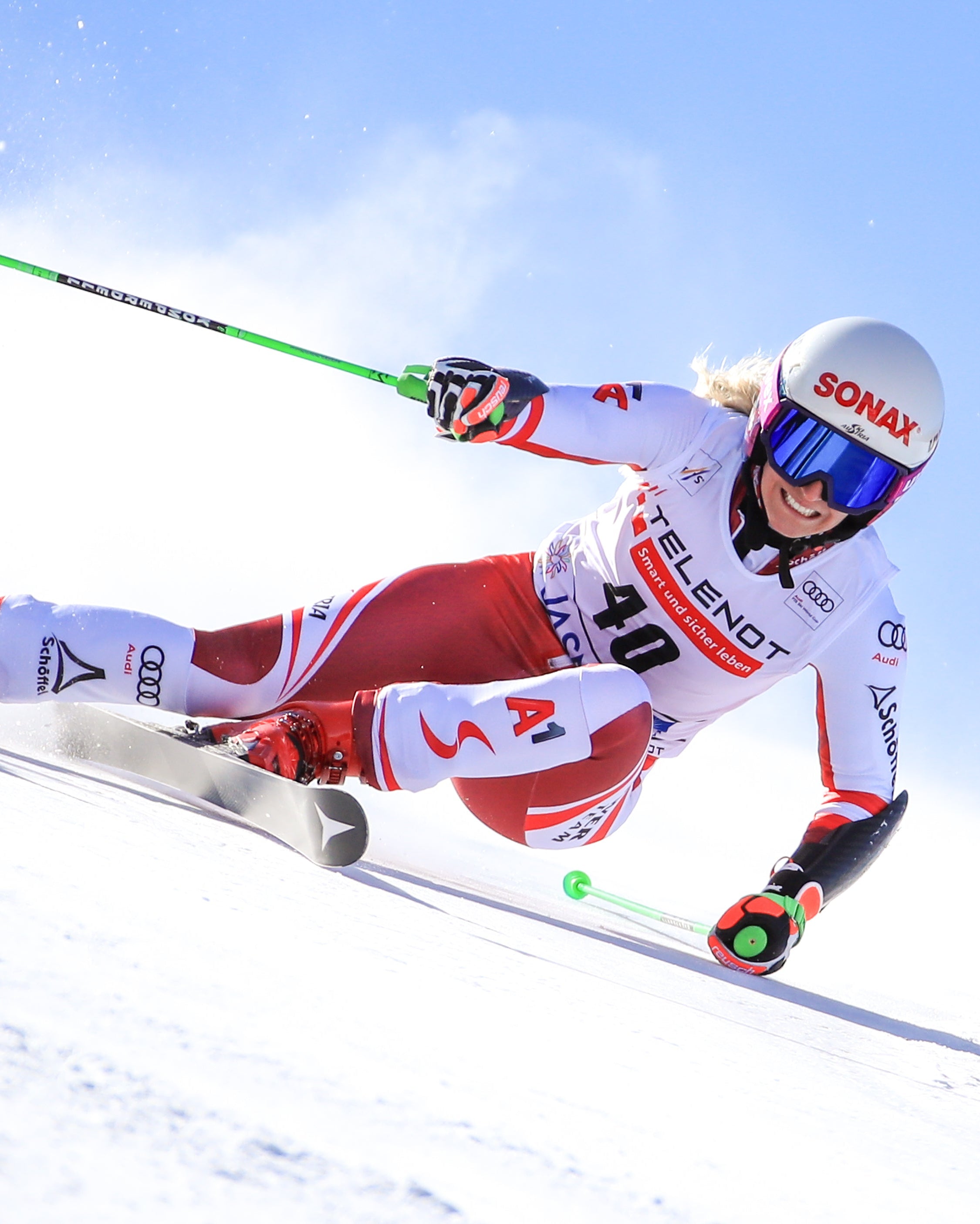 Eva Maria Braem in a red and white racing suit leans sharply into a turn on snow under a clear blue sky, with a red Telenot Jasnau gate flag visible on the right.
