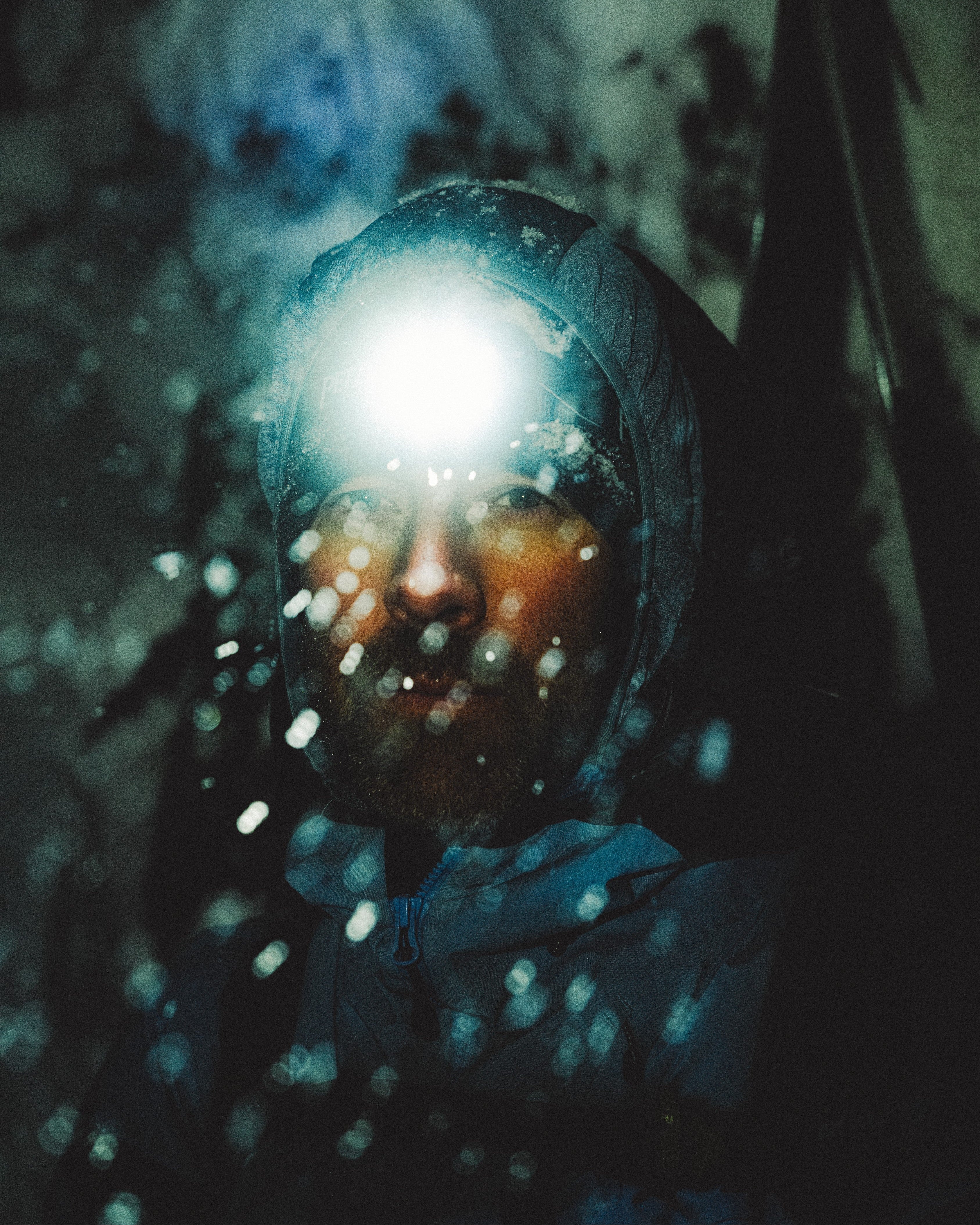 Daron Rahlves wearing a hooded rain jacket with a bright headlamp shining into the camera, with water droplets on the lens and a dark moody outdoor background.