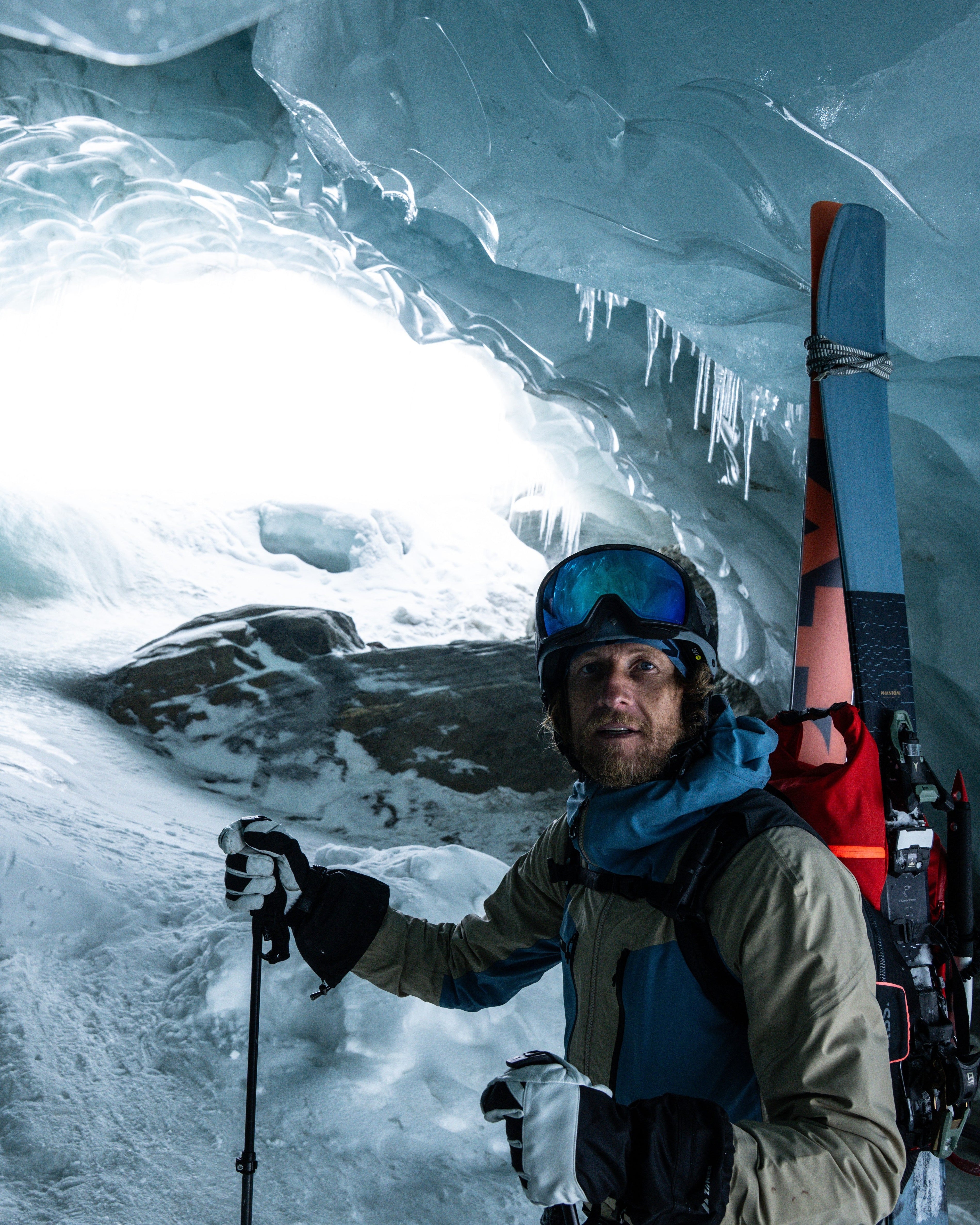 Matthias Haunholder standing inside a glacier cave, wearing ski gear including goggles, a jacket, gloves, and holding a ski pole, with skis strapped to their back.