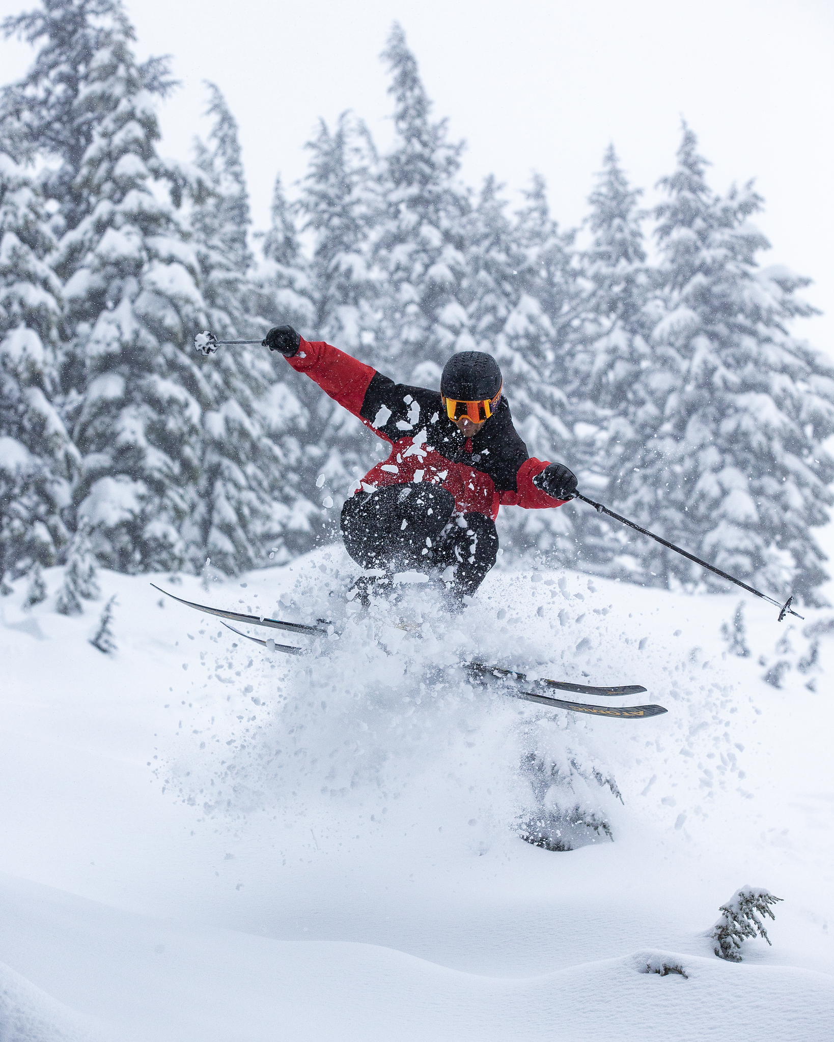 Sage Cattabriga in a red and black jacket, black helmet, and orange-tinted goggles jumps through deep snow, kicking up a spray of powder, with snow-covered pine trees in the background.