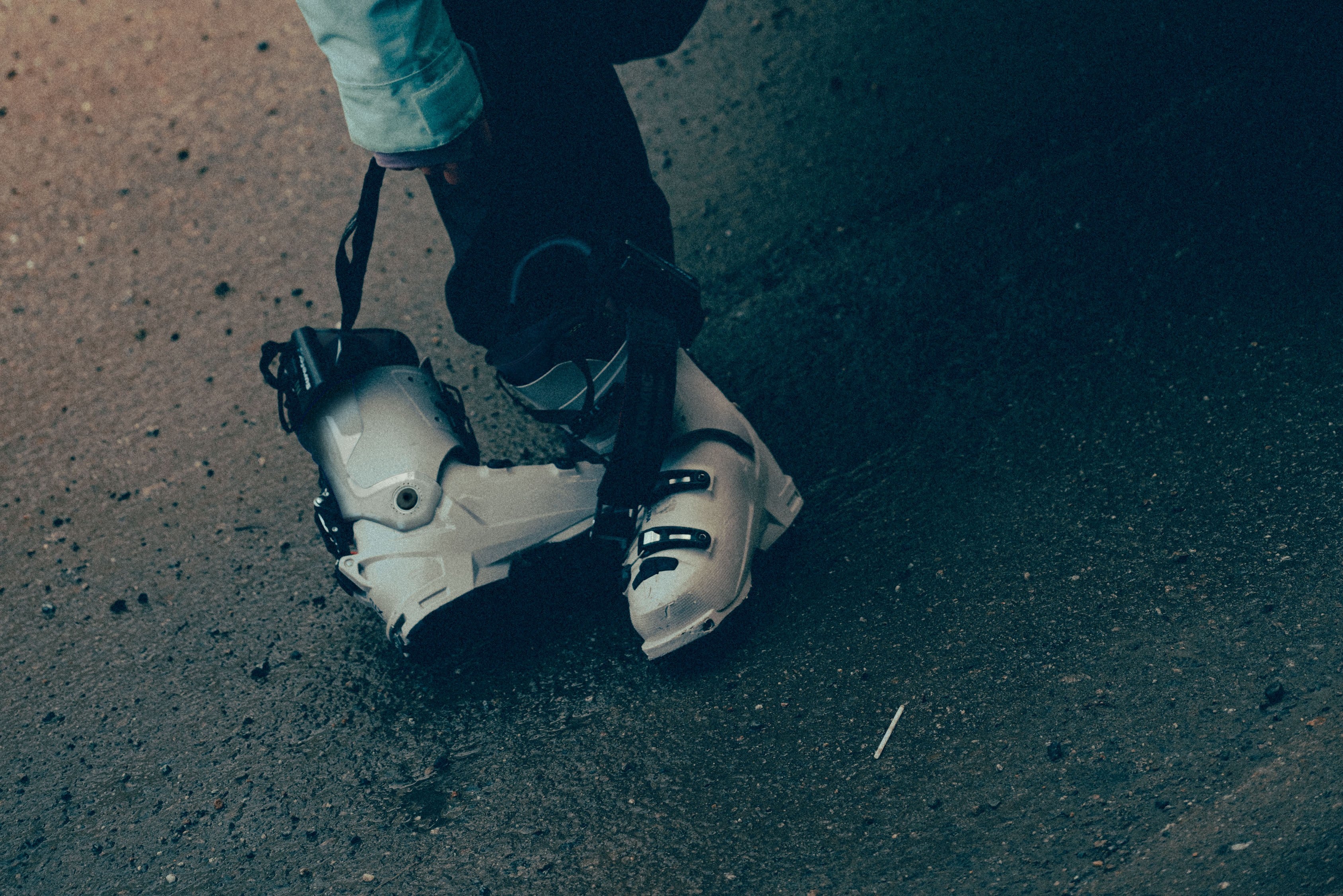 A close-up shot of a person's legs and hands as they buckle a pair Hawx Atomic ski boots on dark asphalt pavement.