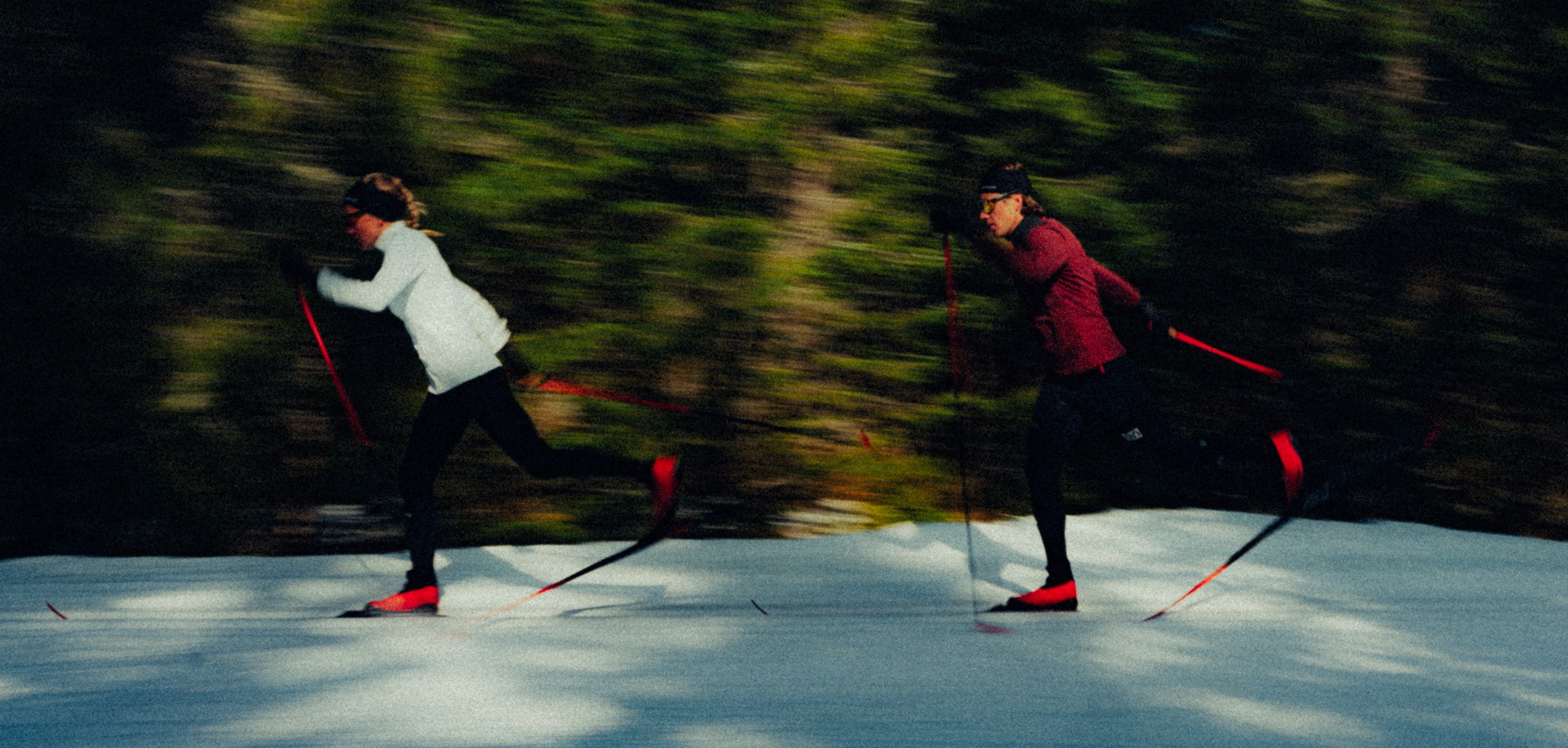 Two people cross-country skiing on snow with a blurred forest background, one wearing a white jacket and the other a dark red jacket.